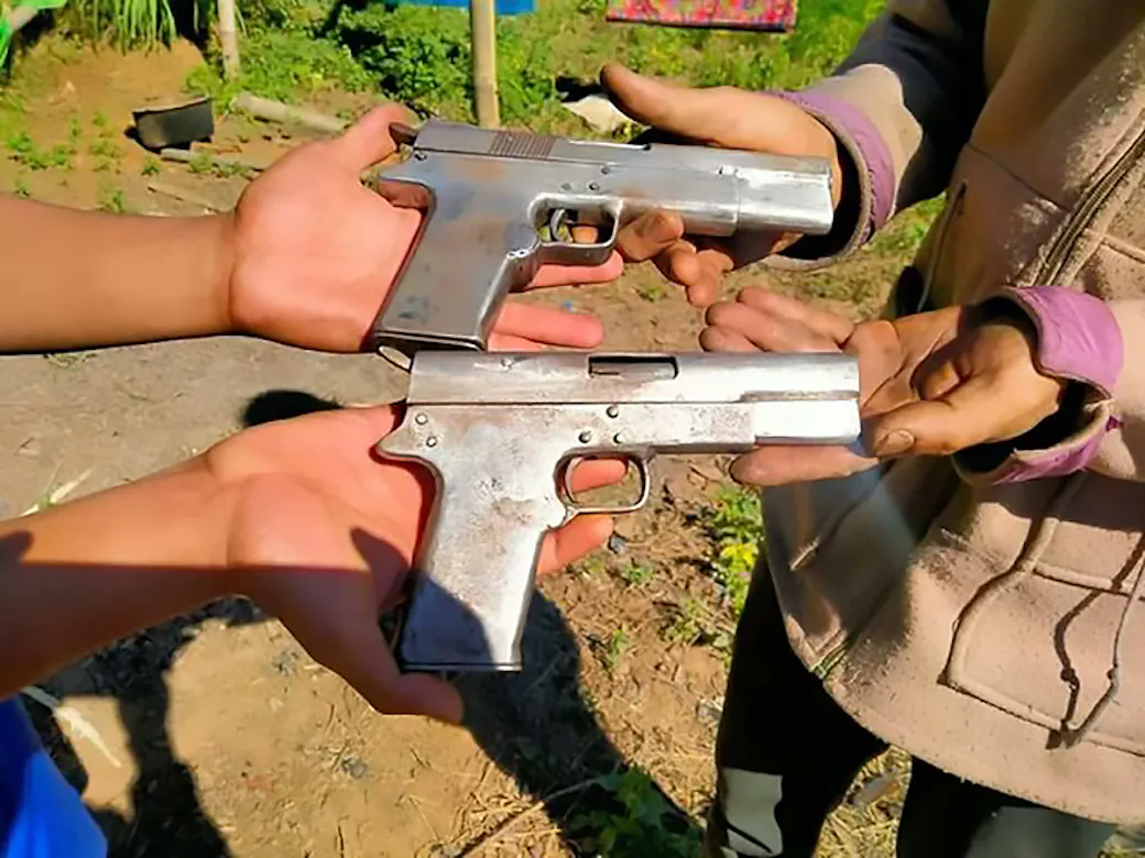 Two crude handmade copies of 1911-style pistols held out in people’s hands in Myanmar