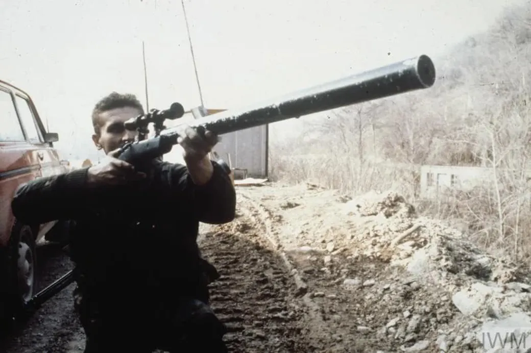 Man aiming a homemade scoped sniper rifle with a long, thick suppressor-style barrel during the Bosnian War